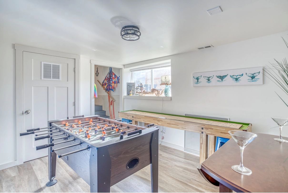 Family entertainment room at Anchor Court Beach House Lincoln City Oregon vacation rental featuring foosball table full size shuffleboard table whale tail coastal wall art beach kites martini glasses on bar table and light hardwood floors — the perfect lower level hangout space for groups and families